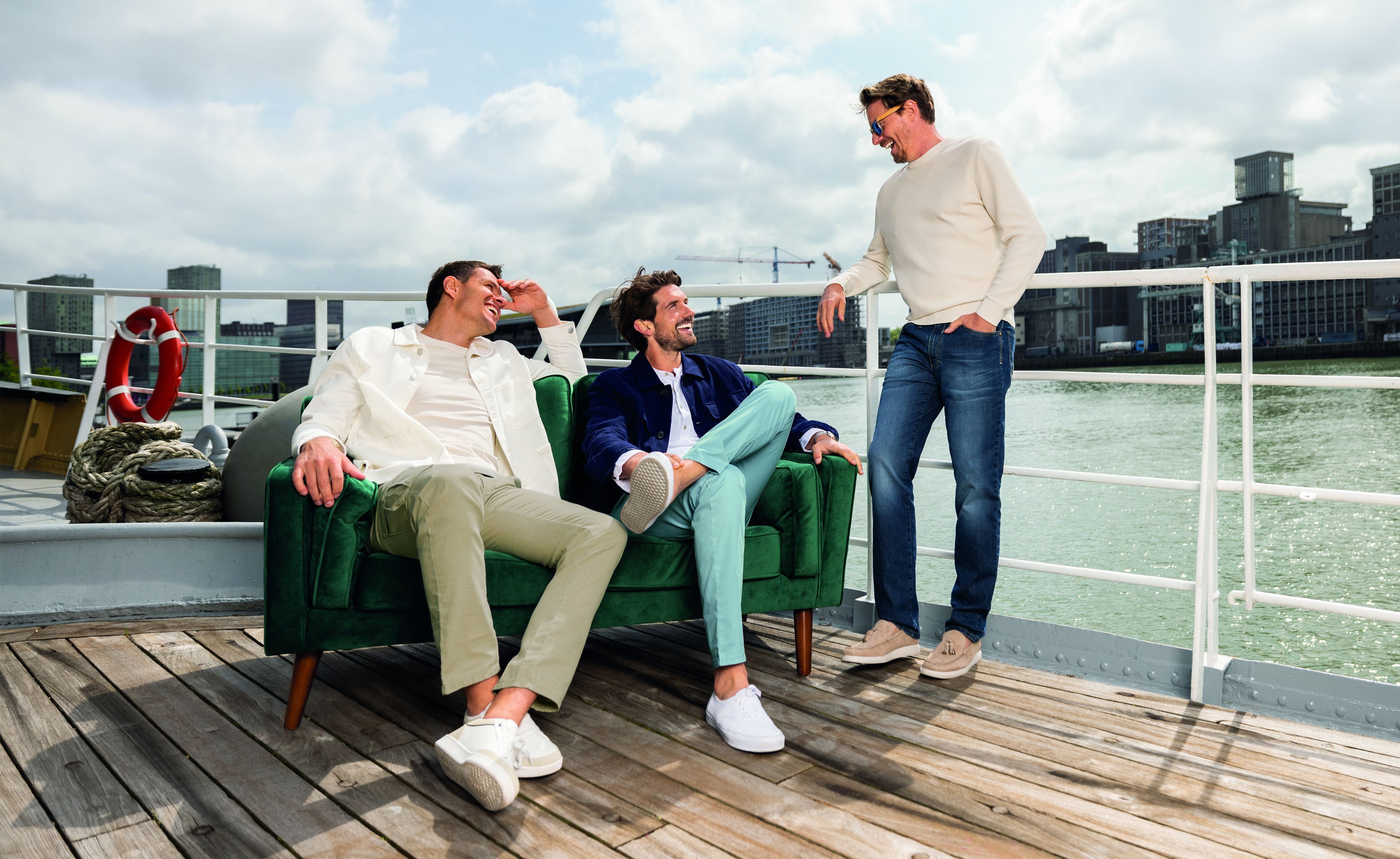 Drei junge Männer sitzen und stehen auf einem Dach mit Blick auf Wasser und Stadt im Hintergrund. Zwei sitzen auf einer grünen Couch, lachen und sprechen, während der dritte Mann im weißen Pullover und Jeans steht, mit einem Fuß auf der Couch, und ihnen freundlich zuspricht. Es ist ein sonniger Tag mit some Wolken am Himmel.