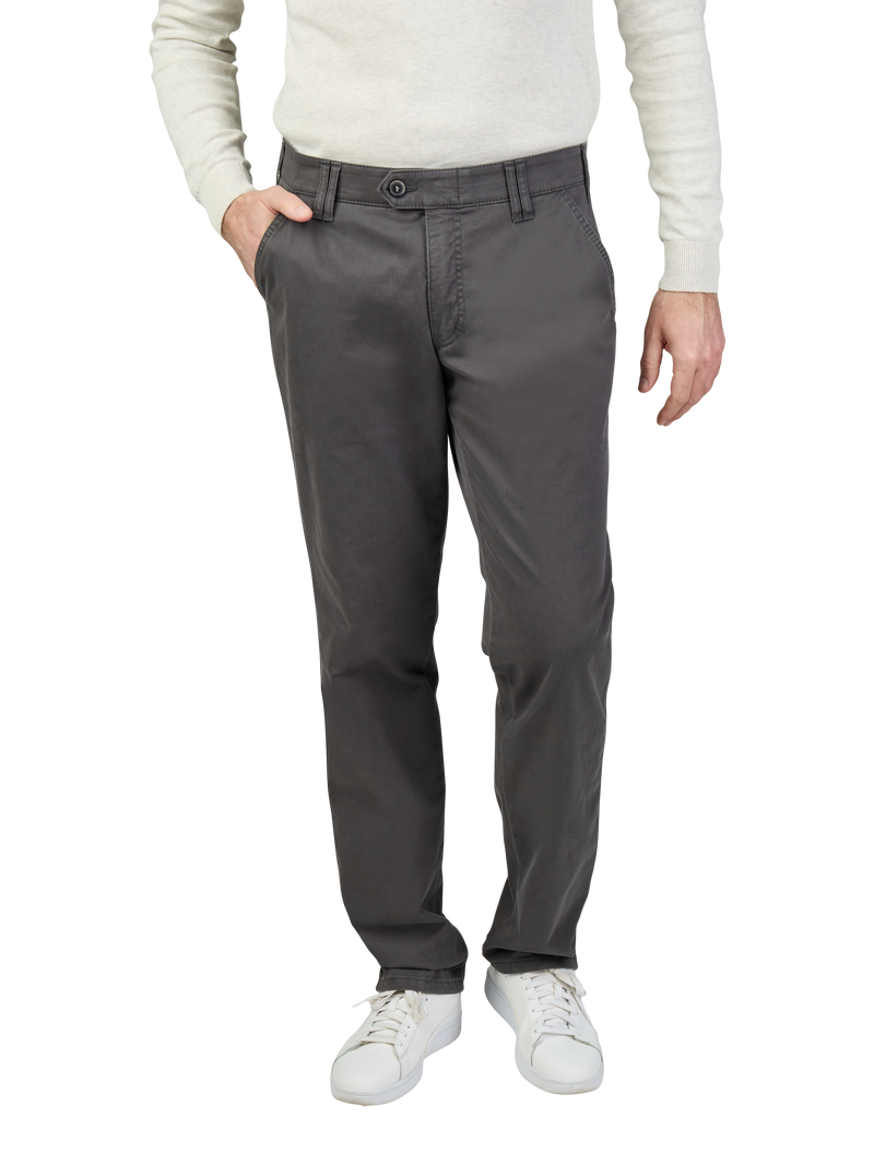 Mann trägt dunkelgraue, gerade geschnittene Chino-Hose mit Gürtelschlaufen und einer Seite mit Hand in der Tasche. Oberkörper trägt er ein hellbeiges, langärmeliges Oberteil, Schuhe sind weiße Sneaker.