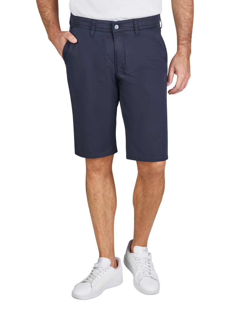 Das Bild zeigt einen Mann, der eine weiße T-Shirt, dunkelblaue Shorts und weiße Turnschuhe trägt. Die Shorts sind knielang, haben Eingrifftaschen an den Seiten und einen Gürtelschlaufen. Der Mann steht mit einer Hand in der Tasche, die andere hängt locker an der Seite.