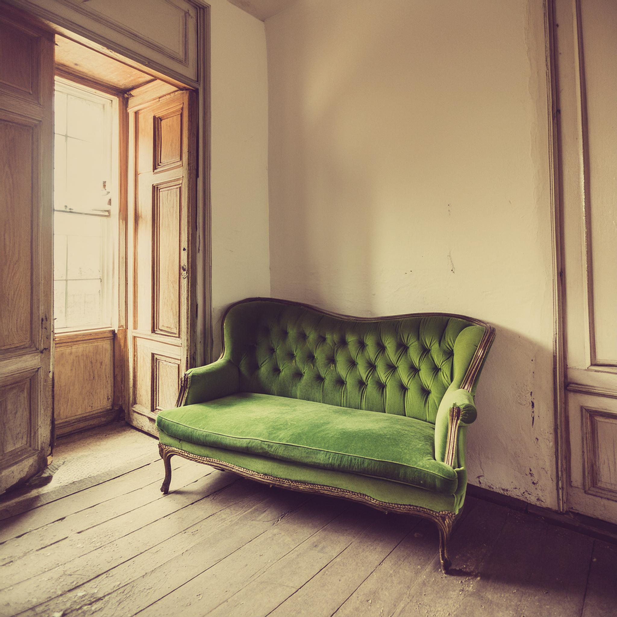 Ein antikes, grünes, samtbezogenes Sofa mit gewölbtem Rücken und geschnitzten Holzbeinen steht in einem Raum mit alten, weißen Wänden und Holz-Doppelfenstern. Die Fensterrahmen und -laden sind rustikal und leicht abgenutzt. Das Zimmer wirkt hell und nostalgisch.