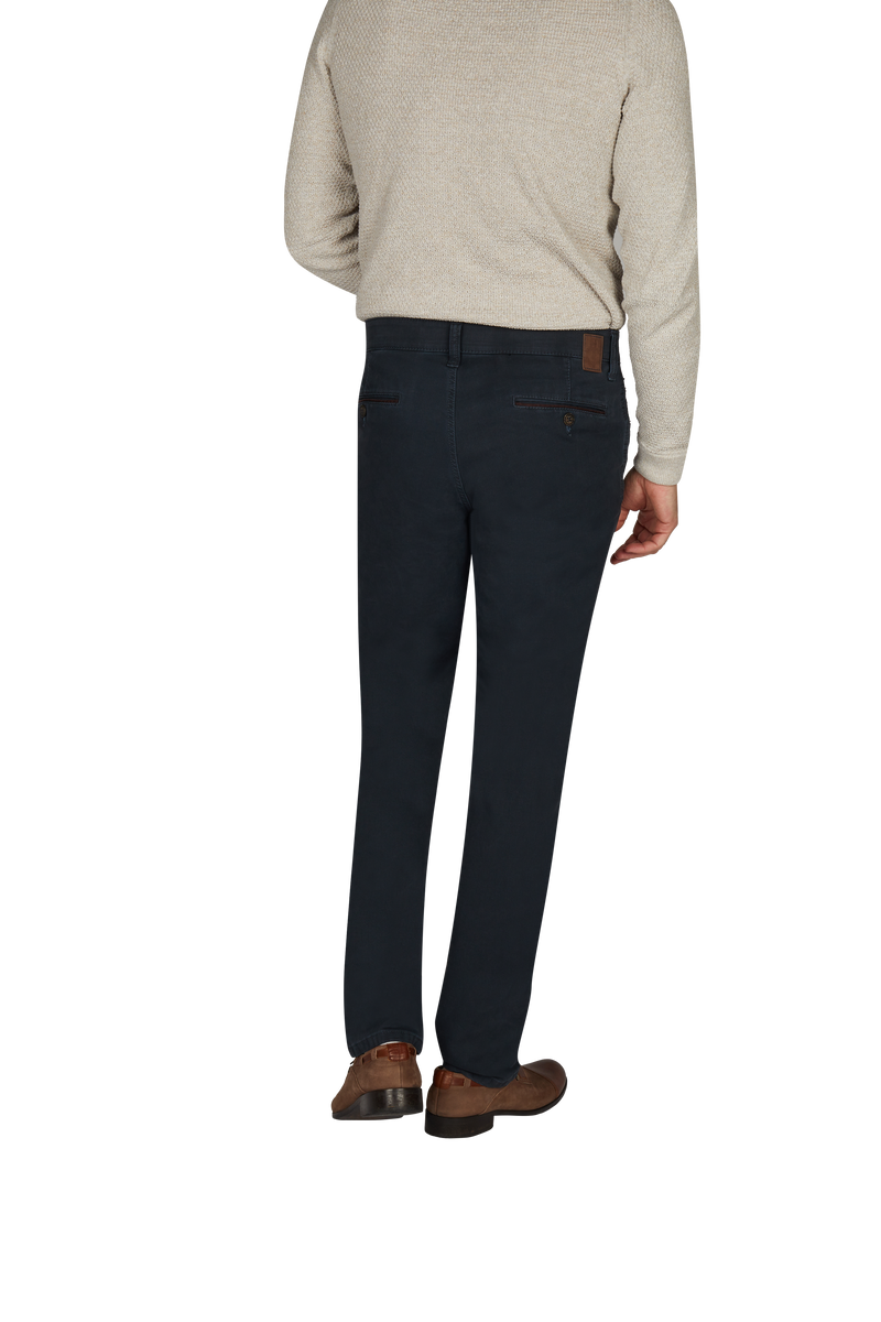 Ein Mann steht mit seinem Rücken zur Kamera, trägt eine beige, langärmlige Pullover und dunkle, lässige Jeans. Er trägt braune Lederschuhe und steht vor einem weißen Hintergrund.