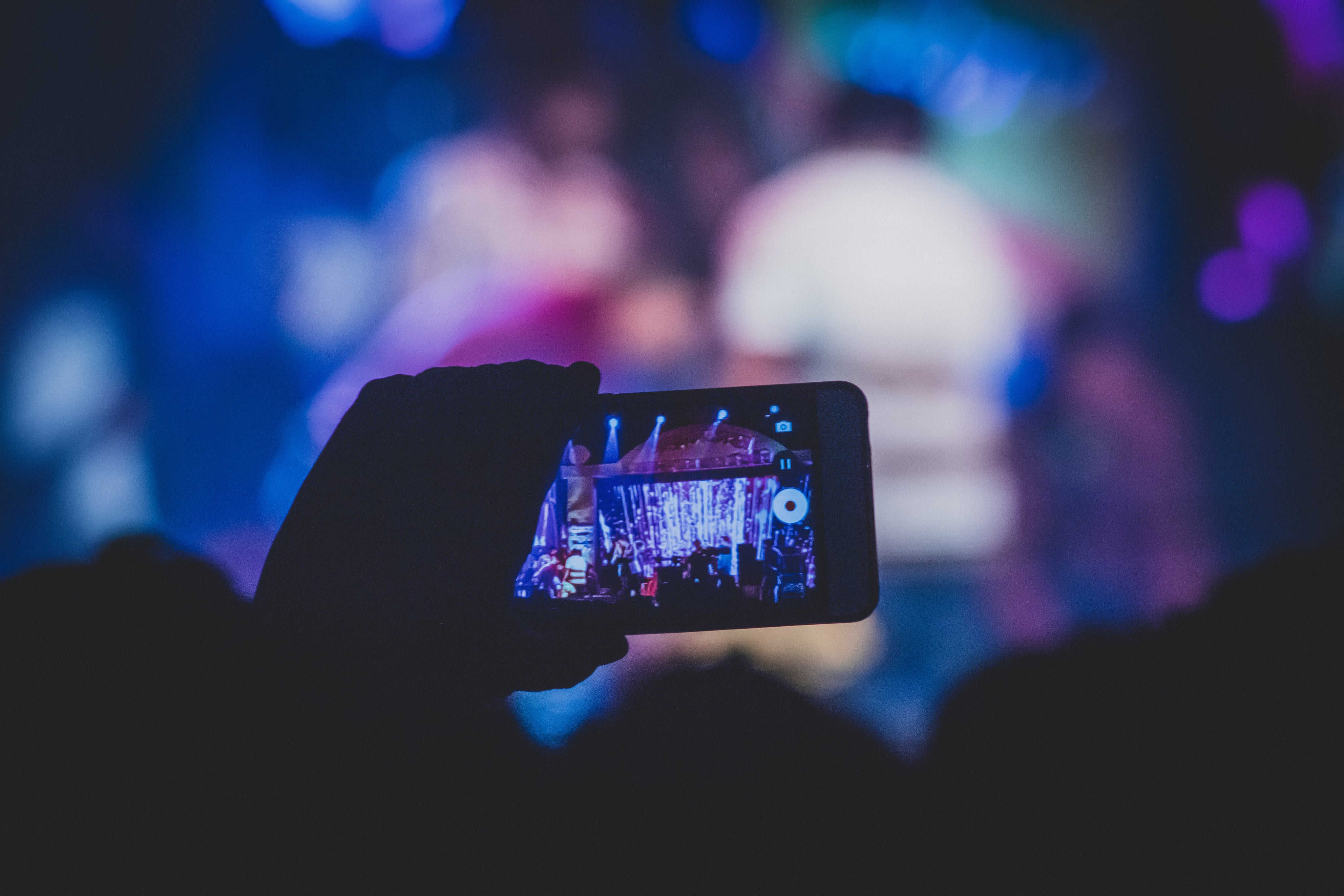 Ein Smartphone wird von einer Person gehalten und zeigt ein Live-Bild eines lebhaften Konzerts mit bunten Lichtern und einer Menschenmenge im Hintergrund, die auf die Bühne schaut.