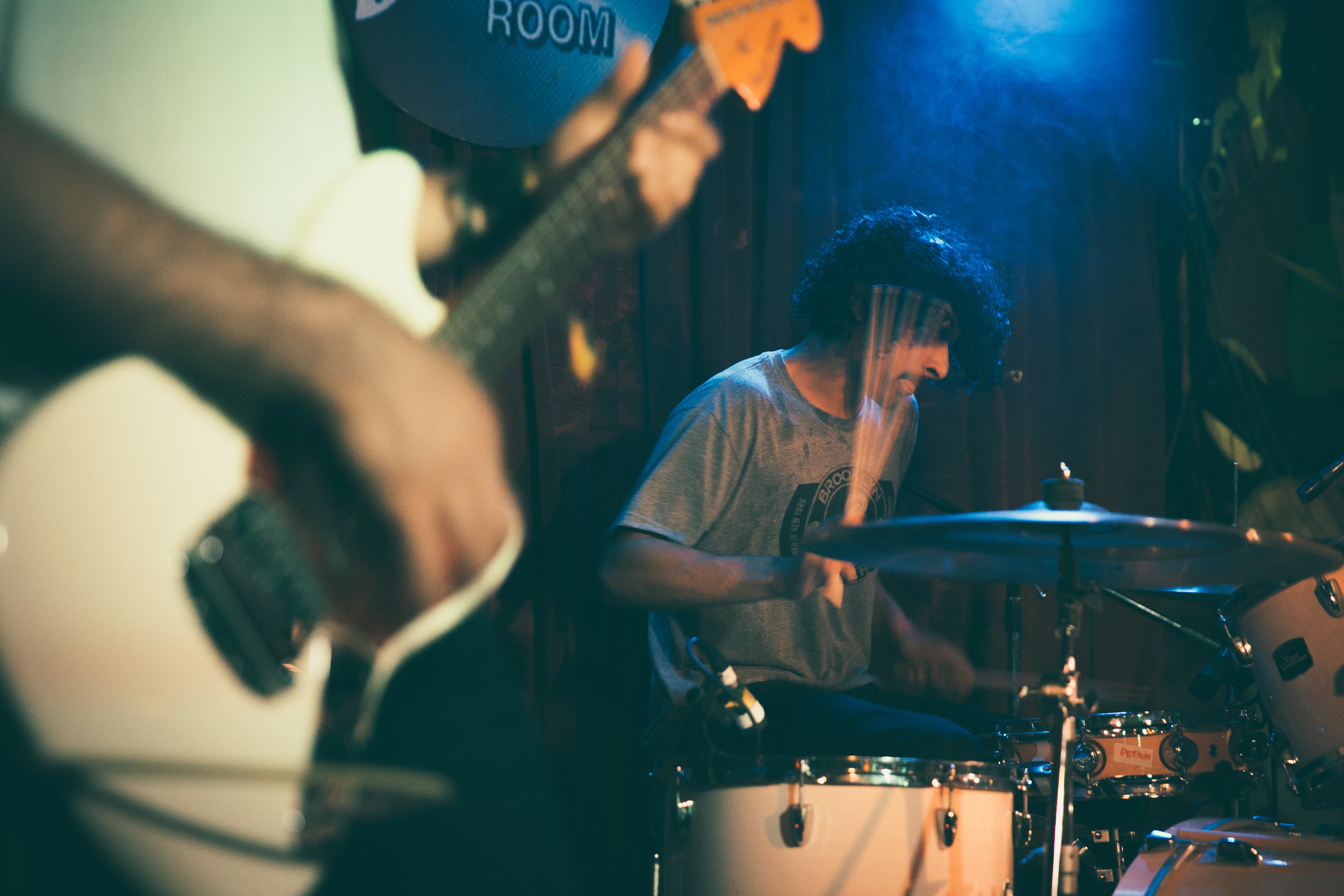 Ein junger Musiker mit curly Haaren spielt auf einem Drum-Set in einer dunklen, mit blauen Lichtern beleuchteten Bühne. Im Vordergrund ist die Hand eines anderen Musikers mit einer Gitarre sichtbar.