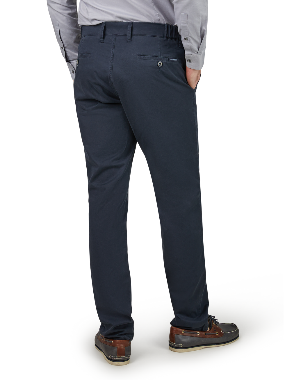 Ein Mann trägt schwarze Chino-Hosen, eine graue Hemd und braune Schuhe. Er steht mit den Händen in den Taschen, zeigt den Rücken zum Betrachter.